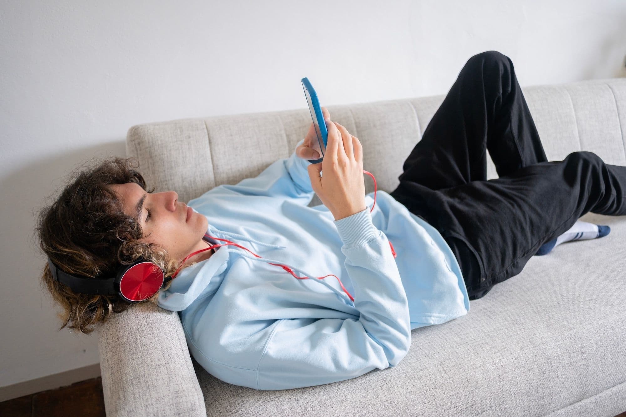 A person with curly hair wearing a light blue hoodie and red headphones lies on a light-colored sofa, looking at a tablet or phone.