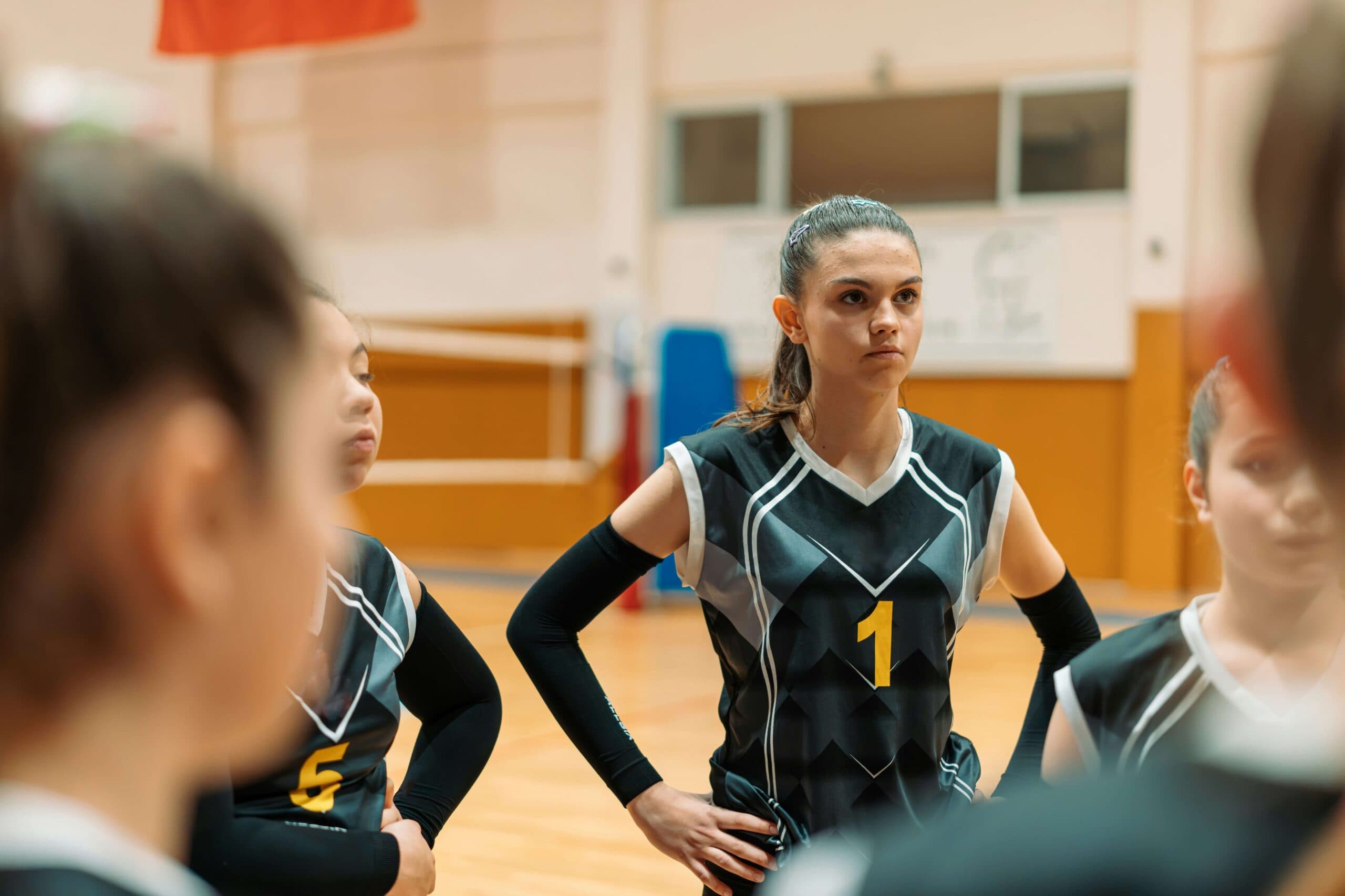 A volleyball player wearing a black jersey with the number 1 stands with teammates in a gym, arms on hips, appearing focused and ready to cope with sports pressure.